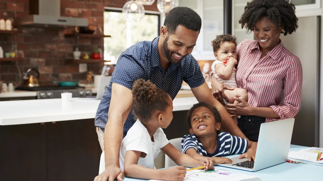A family of five is in the kitchen, helping two of their children with their homework.