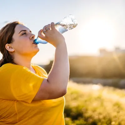 A woman drinking a bottle of water on a sunny day, surrounded by yellow flowers.
