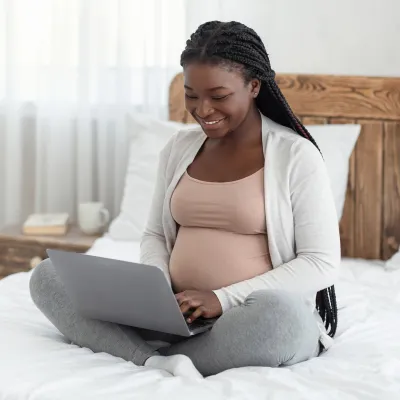 A Smiling Pregnant Woman Sits on Her Bed, in Her Home, Using a Laptop 