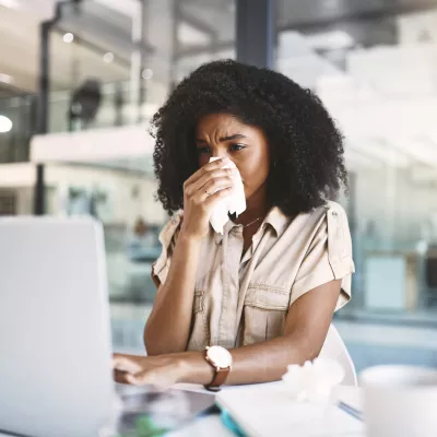 A woman blowing her nose, looking at a laptop.