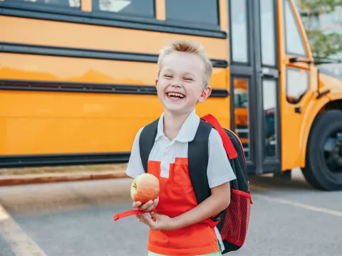 A Small Boy Laughs as He Gets Off a School Bus with an Apple in Hand.