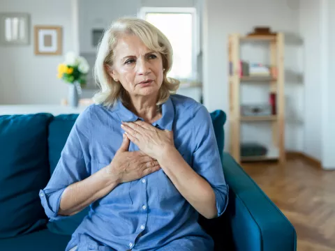 A Woman Sits in Her Living Room With Her Hands on Her Chest, Feeling Her Heartbeat.
