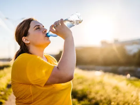 A woman drinking a bottle of water on a sunny day, surrounded by yellow flowers.
