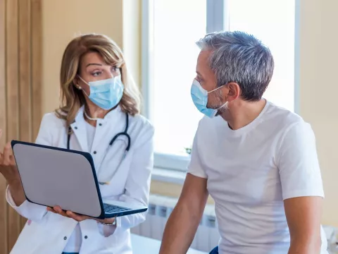 A Doctor Speaks to Her Patient in an Exam Room While Going Over His Charts on a Laptop.