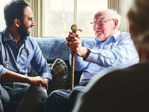 A doctor, sitting on a sofa with a senior man holding a cane, making a home visit.