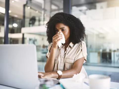 A woman blowing her nose, looking at a laptop.