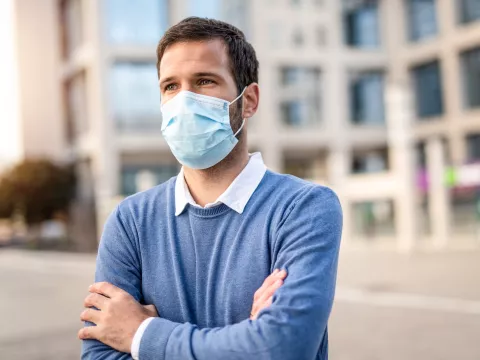 A man with his arms crossed standing outdoors wearing a mask.